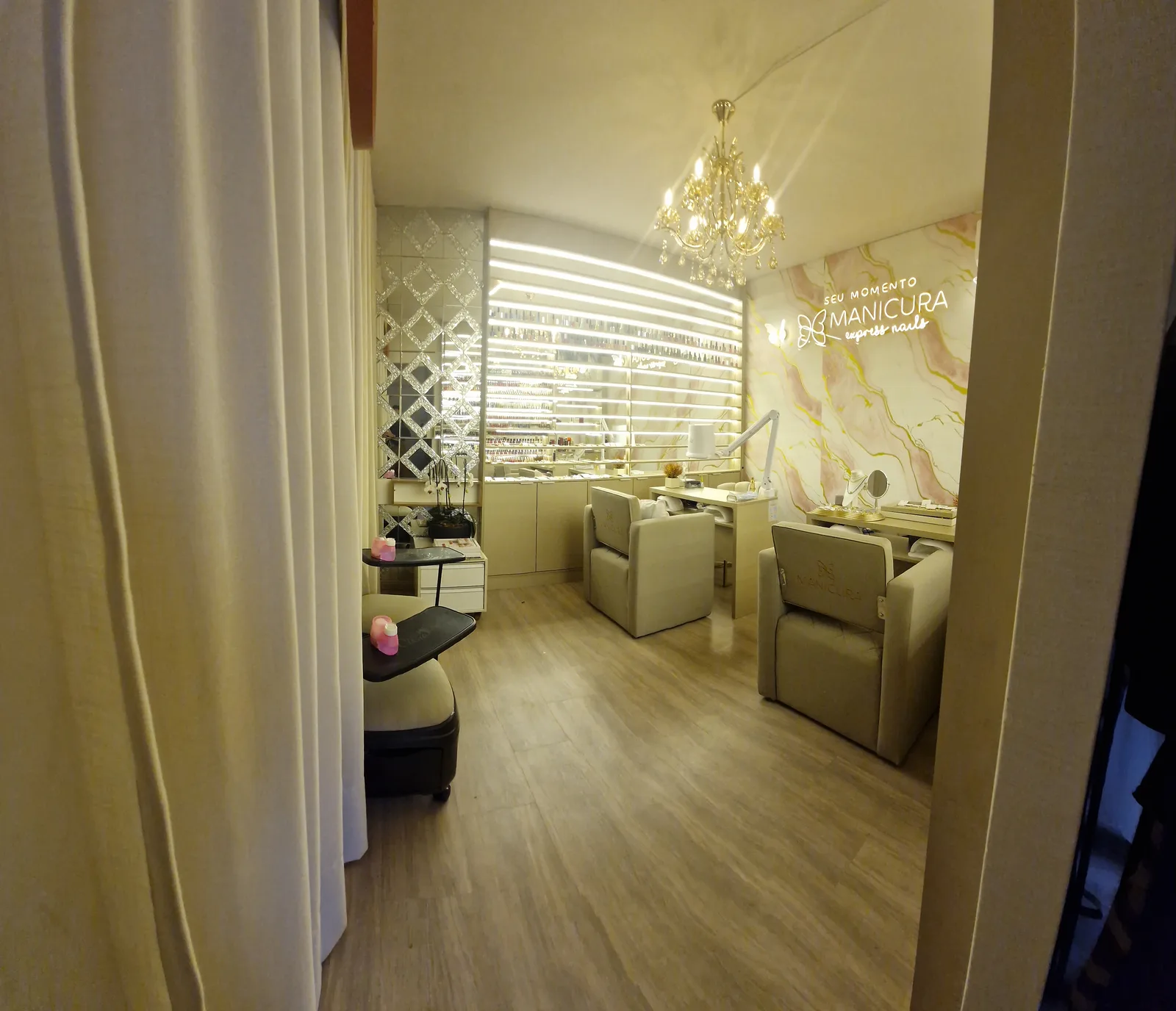 Estúdio Manicura Express Nails visto da entrada, entre as cortinas, com estações de atendimento, lustre e letreiro ao fundo