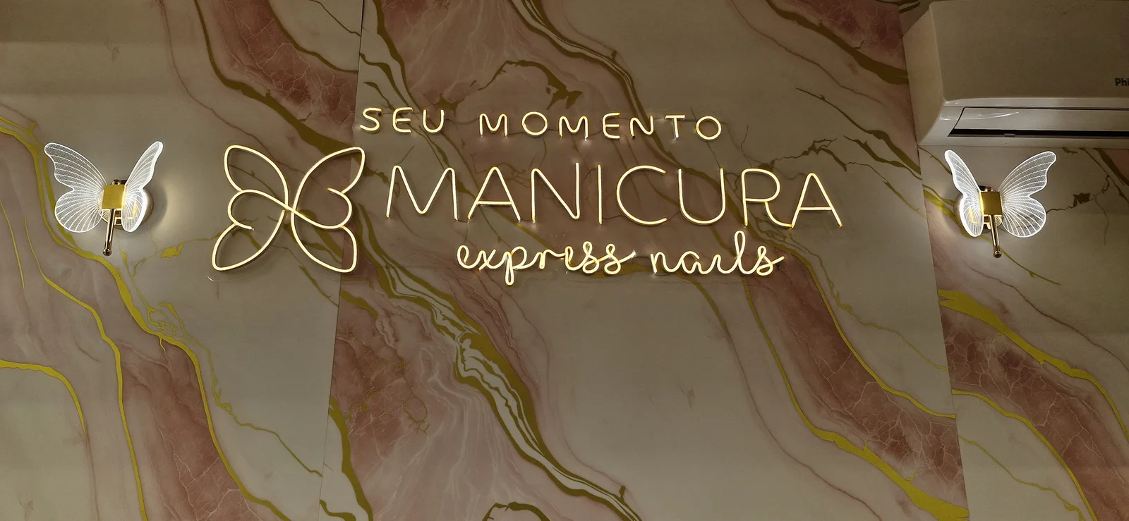 Letreiro iluminado SEU MOMENTO Manicura Express Nails com arandelas de borboleta na parede de mármore do estúdio